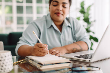 Frau sitzt am Schreibtisch und schreibt Notizen in ein Tagebuch. Ausschnitt einer Frau, die sich Notizen macht, während sie zu Hause am Schreibtisch arbeitet. - JLPSF03545