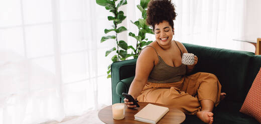 Beautiful woman sitting on sofa with coffee mug and checking mobile phone. Female using smart phone and holding coffee mug sitting in living room. - JLPSF03550