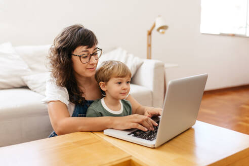 Mutter und Sohn benutzen einen Laptop zu Hause. Eine Frau benutzt einen Laptop und ihr Sohn schaut zu, während sie im Wohnzimmer sitzt. - JLPSF03558