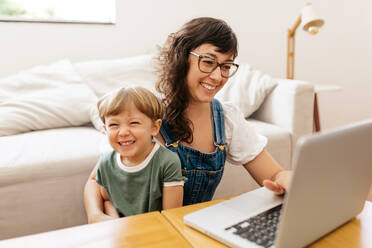 Woman working on laptop with her son sitting on her lap and smiling. Happy mother and son at home. - JLPSF03559