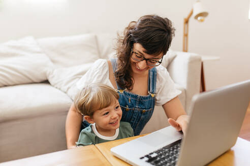 Kleiner Junge schaut auf den Laptop, während er mit seiner Mutter zu Hause sitzt. Mutter und Sohn schauen sich etwas Interessantes auf dem Laptop im Wohnzimmer an und lächeln. - JLPSF03560