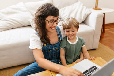 Frau mit ihrem Sohn, der an ihrem Laptop sitzt und diesen zu Hause benutzt. Arbeitende Mutter, die sich um ihren Sohn kümmert und zu Hause einen Laptop benutzt. - JLPSF03561