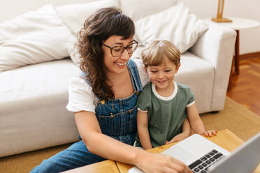 Woman with her son sitting on her laptop using laptop at home. Working mother taking care of her son and using laptop at home. - JLPSF03561