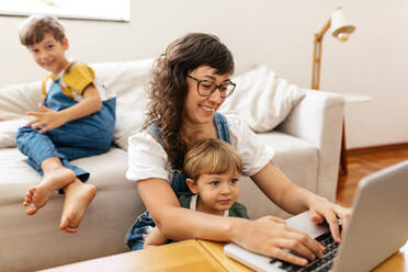 Frau arbeitet am Laptop mit ihrem Sohn sitzt auf ihrem Schoß und anderen Sohn sitzt auf dem Sofa im Hintergrund. Mutter suchen beschäftigt Arbeit am Laptop mit Kindern zu Hause. - JLPSF03562