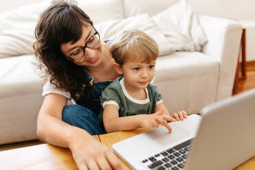 Kleiner Junge benutzt Laptop, während er mit seiner Mutter zu Hause sitzt. Kind lernt mit seiner Mutter im Wohnzimmer den Umgang mit dem Laptop. - JLPSF03564