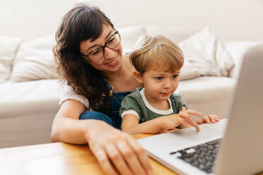 Cute boy using laptop trackpad while sitting with his mother at home. Child learning to use laptop with his mother. - JLPSF03565