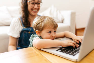 Ein Junge tippt auf einem Laptop, während seine Mutter im Hintergrund sitzt. Ein Kind benutzt einen Laptop, während seine Mutter neben ihm sitzt und lächelt. - JLPSF03566