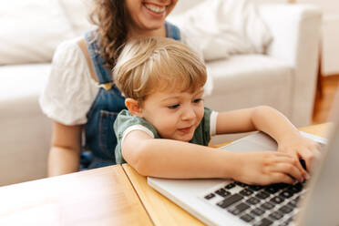 Kid pressing keys of laptop keyboard with mother sitting by. Boy learning using laptop with mother at home. - JLPSF03567