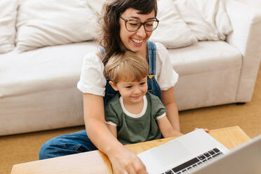 Mutter und Sohn schauen gemeinsam auf den Laptop und lächeln. Frau benutzt Laptop mit ihrem Sohn, der zu Hause auf ihrem Schoß sitzt. - JLPSF03568