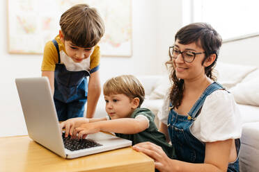 Ein Junge benutzt einen Laptop, während seine Mutter und sein Bruder daneben stehen. Eine Mutter mit zwei Kindern benutzt einen Laptop im Wohnzimmer. - JLPSF03571