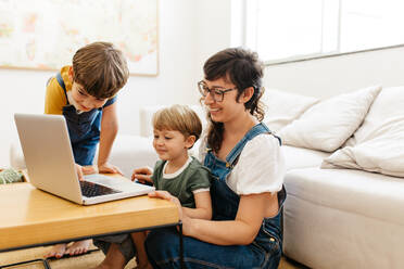 Niedliche junge Familie, die zu Hause gemeinsam auf einen Laptop schaut. Mutter sitzt mit ihren beiden Jungen im Wohnzimmer und schaut auf den Laptop auf dem Tisch. - JLPSF03572