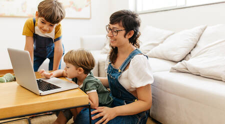 Kinder, die mit ihrer Mutter zu Hause den Umgang mit dem Laptop lernen: Ein Junge, der auf dem Schoß seiner Mutter sitzt und sein Bruder, der im Wohnzimmer steht. - JLPSF03573