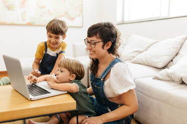 Mutter und ihre Kinder benutzen einen Laptop zu Hause. Eine dreiköpfige Familie benutzt einen Laptop, während sie im Wohnzimmer sitzt. - JLPSF03574