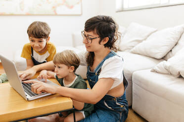 Mutter zeigt ihren Kindern etwas Interessantes auf dem Laptop. Mutter und Jungen schauen sich etwas Interessantes auf dem Laptop im Wohnzimmer an. - JLPSF03576