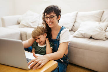 Woman working on laptop with her son sitting on her lap. Mother sitting on floor working from home. - JLPSF03579