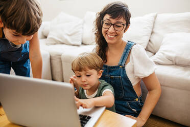 Lächelnde Frau, die ihren Sohn beim Lernen mit dem Laptop beobachtet, während sein älterer Sohn daneben steht. Kleine glückliche Familie mit Laptop zu Hause. - JLPSF03584