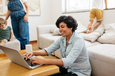 Happy woman working on laptop with her family in background. Female using laptop in front with her family at home. - JLPSF03590