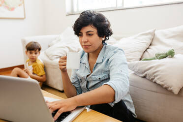 Woman drinking coffee while working on laptop at home. Mother busy working on laptop with her son sitting in background. - JLPSF03593