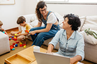 Glückliches lesbisches Paar zu Hause mit Kindern. Eine arbeitet am Laptop und die andere spielt mit den Kindern. Beide schauen sich an und lächeln. - JLPSF03596