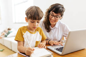 Ein Junge schreibt an einem Buch, während seine Mutter am Laptop arbeitet. Ein Kind lernt mit seiner Mutter, die von zu Hause aus arbeitet. - JLPSF03604