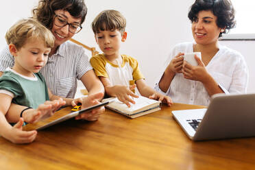 Woman using digital tablet while sitting with her family at home. Lesbian couple spending some time with their two sons at home. - JLPSF03609