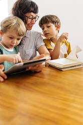 Niedlicher Junge, der ein digitales Tablet benutzt, während er mit Mutter und Bruder zusammensitzt. Junge Familie zusammen mit einem digitalen Tablet in einem Haus. - JLPSF03610