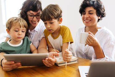 Eine Familie sitzt zusammen am Tisch und benutzt ein digitales Tablet. Eine Frau mit Kindern schaut auf einen Laptop, während ihr Partner Kaffee trinkt und lächelt. - JLPSF03611