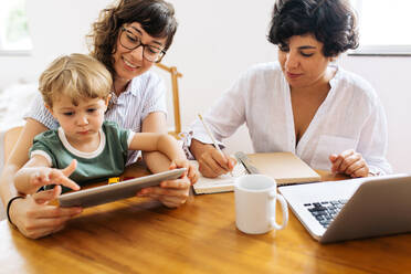 LGBT couple at home with kid. One working and another sitting by with son using digital tablet. - JLPSF03612