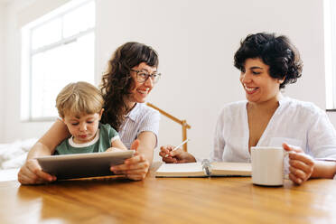 Lesbian couple looking at each other and smiling with son using tablet pc at home. Female couple smiling while their son using digital tablet. - JLPSF03613