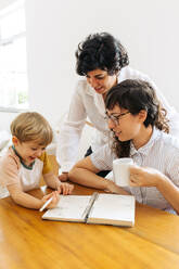 Ein Junge hat Spaß, während er mit seinen Müttern zu Hause das Schreiben lernt. Ein weibliches Paar trinkt Kaffee mit seinem Sohn, der am Tisch sitzt und an einem Buch schreibt und lächelt. - JLPSF03617