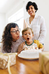 Lesbische Eltern feiern den Geburtstag ihres Sohnes zu Hause. Junge mit Mutter bläst die Kerzen auf dem Geburtstagskuchen aus. - JLPSF03621