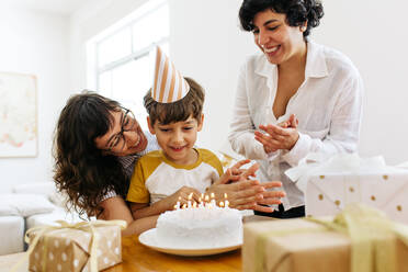 Lächelnde Mütter mit Sohn, die den Kuchen mit angezündeten Kerzen betrachten und in die Hände klatschen. Lesbische Eltern feiern den Geburtstag eines Jungen. - JLPSF03624