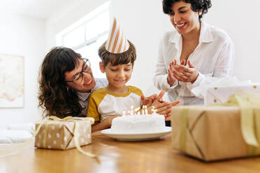 Lächelnde Mütter mit ihrem Sohn betrachten einen Kuchen mit brennenden Kerzen. Homosexuelle Eltern feiern den Geburtstag eines Jungen. - JLPSF03625