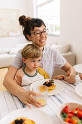Mutter und Sohn beim gemeinsamen Frühstück. Lächelnde Frau mit ihrem Sohn auf dem Schoß beim Frühstück zu Hause. - JLPSF03636