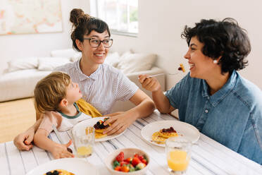 Weibliches Paar mit Sohn beim Frühstück zu Hause: Lächelnde Frauen, die sich beim Frühstück ansehen, während ihr Sohn zuschaut. - JLPSF03637