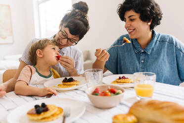 Eine Frau füttert ihren Sohn beim Frühstück mit Obst. Ein lesbisches Paar frühstückt mit seinem Sohn zu Hause. - JLPSF03638