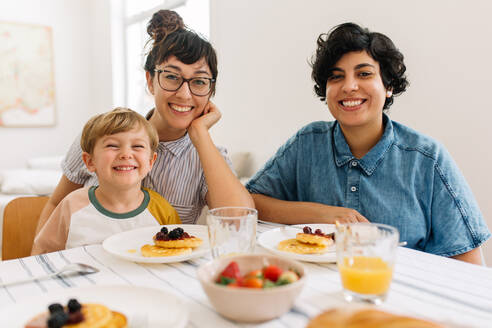 Porträt einer schönen dreiköpfigen Familie, die am Frühstückstisch sitzt, in die Kamera schaut und lächelt. Lesbisches Paar mit Sohn beim gemeinsamen Frühstück. - JLPSF03639