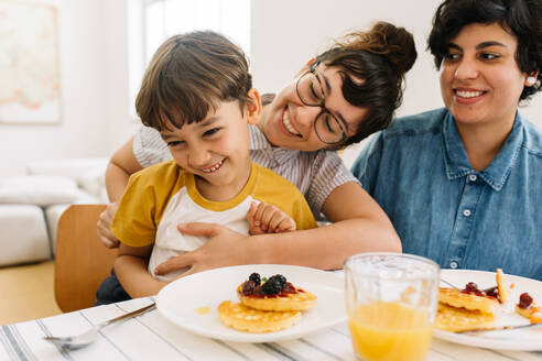 Familie, die sich beim Frühstück amüsiert. LGBT-Paar sitzt lächelnd am Frühstückstisch, wobei die Frau ihren Sohn kitzelt. - JLPSF03640
