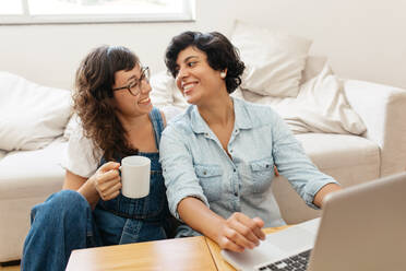 Female couple sitting together at home. Woman holding coffee cup and looking at her partner working on laptop in living room. - JLPSF03651