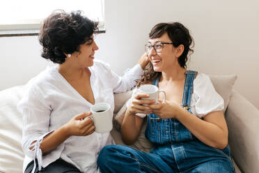Liebevolles Frauenpaar, das zu Hause zusammen sitzt und Kaffee trinkt. Gleichgeschlechtliches Frauenpaar, das zusammen auf dem Sofa sitzt und Kaffee trinkt und sich unterhält. - JLPSF03654