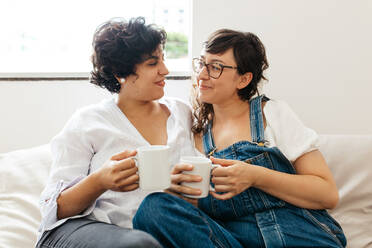 Lesbisches Paar, das sich beim Kaffeetrinken auf dem Sofa im Wohnzimmer liebevoll anschaut. Weibliches Paar, das süße Momente miteinander teilt. - JLPSF03655
