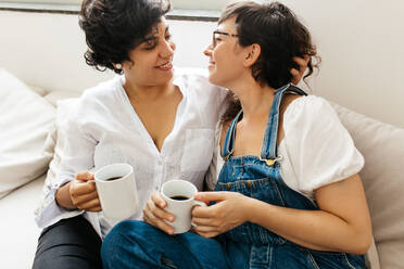 Lesbisches Paar mit Kaffee in der Hand, das sich küssen will. Verliebte Frauen sitzen zusammen auf dem Sofa zu Hause. - JLPSF03656