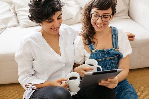 Glückliches lesbisches Paar, das Kaffee trinkt und ein digitales Tablet im Wohnzimmer benutzt. Schönes Frauenpaar zu Hause mit einem Tablet-Computer. - JLPSF03657