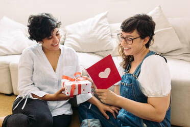 Frau gibt ein Geschenk und liest eine Karte für ihre Freundin zu Hause. Lesbisches Paar mit Geschenk und Valentinskarte. - JLPSF03667