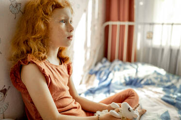 Nachdenkliches rothaariges Mädchen mit Stofftier auf dem Bett sitzend - ANAF00108