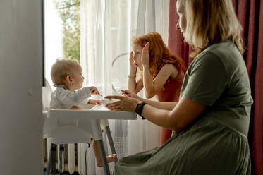 Woman feeding baby girl playing with sister at home - ANAF00114