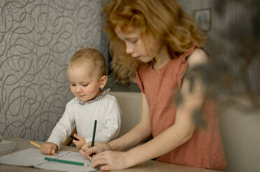Girl teaching sister to draw in book at home - ANAF00120
