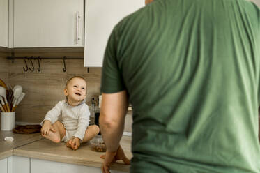 Happy cute baby girl sitting on counter looking at father standing in kitchen - ANAF00139