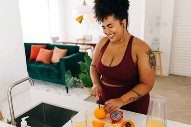 Portrait of healthy woman standing and cutting fruits at home in kitchen. Plus size female in exercise outfit preparing juice at home. - JLPSF03721