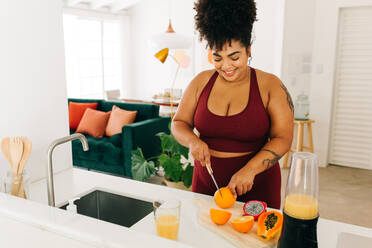 Body positive woman cutting fruits while standing in kitchen. Healthy female in fitness wear preparing juice at home. - JLPSF03722